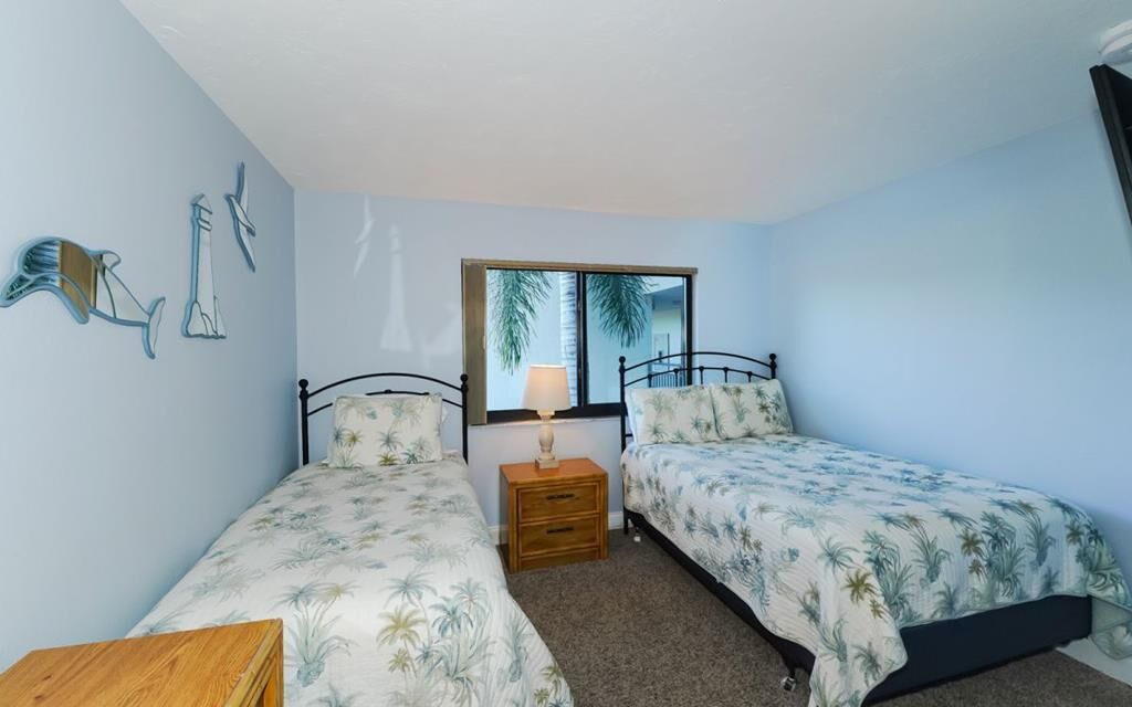Une chambre avec deux lits simples, une table de chevet avec une lampe, une table en bois et une fenêtre donnant sur des palmiers.