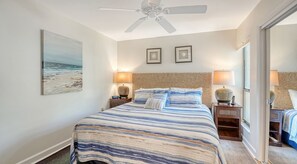 A bedroom with a bed, two bedside lamps, a ceiling fan, and a beach painting on the wall.