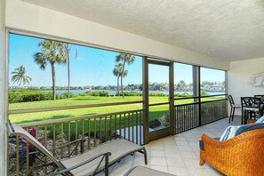 A balcony with a view of a lake and palm trees, furnished with a sofa and chairs. - Chinaberry 913-Recently refurnished and re-decorated! Ground-level, bay front, 2BR/2BA that sleeps 6 (Siesta Key)