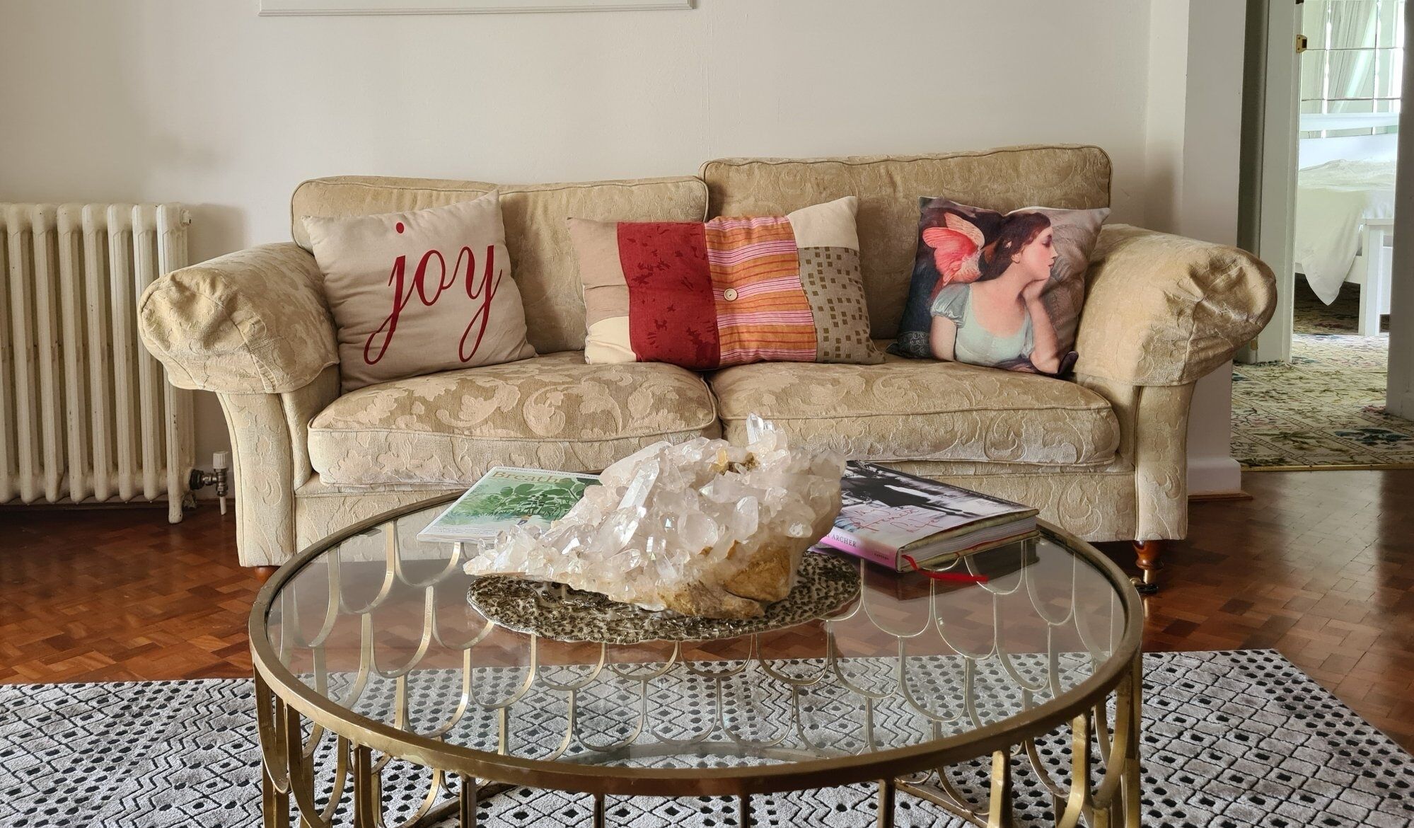 Una sala de estar con un sofá color crema decorado con varios cojines, una mesa de centro de vidrio con un centro de cristal, y un radiador al fondo.