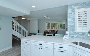 A kitchen with white cabinetry, a central island, and a dining area with teal chairs. - Doveplum 7224- 2BR canal-view townhouse in Sarasota with pool, WiFi, and private beach access. (Siesta Key)