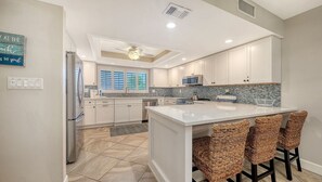A modern kitchen with white cabinets, a central island with bar stools, a built-in oven, and a window with blinds.