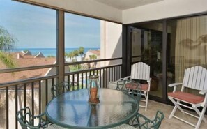 A balcony with a glass table and chairs, overlooking a beach and ocean view.