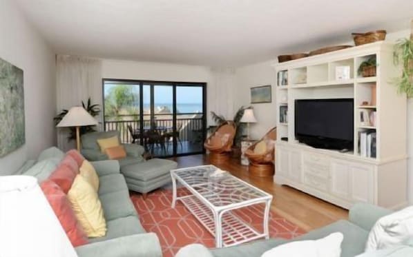 A living room with a sofa, a coffee table, and a television. There is a balcony with a view of the sea.