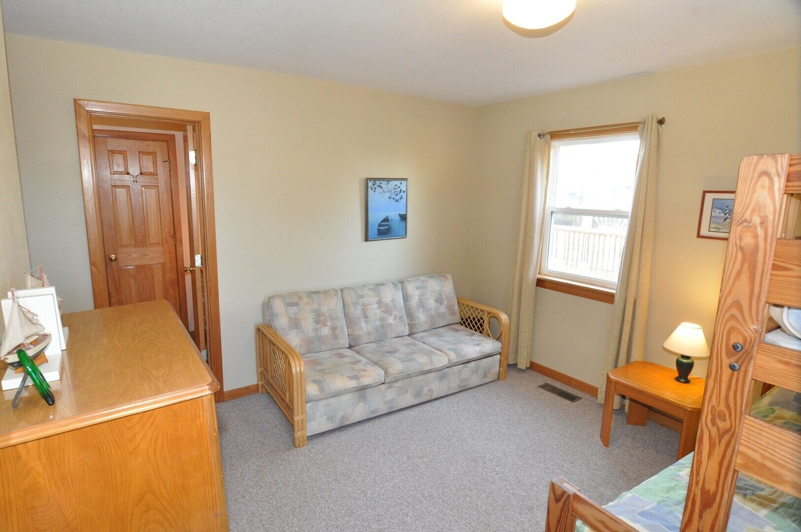 A room with a wooden desk, a sofa, a small table with a lamp, and a window with curtains.