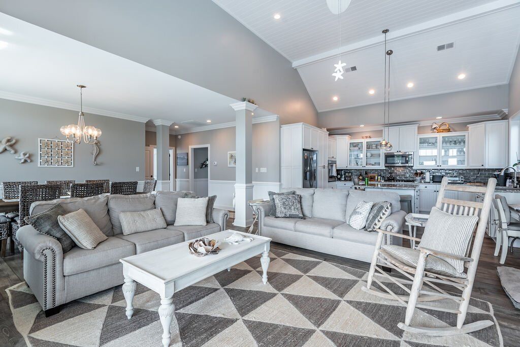 A spacious living room with a large sofa, a coffee table, and a rocking chair. The room has a high ceiling with recessed lighting and a chandelier. There is a kitchen area visible in the background with white cabinets and a countertop.
