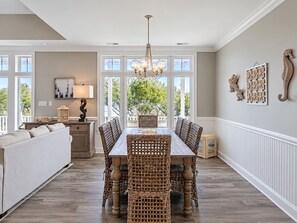 Un coin repas avec une table en bois et des chaises en osier, un lustre et une vue sur l’extérieur grâce à de grandes fenêtres.