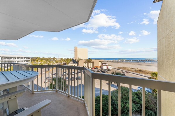 A balcony with a view of a beach, parking area, and a building in the background.