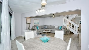 Un salon moderne avec une table ronde en bois, des chaises blanches, un canapé gris agrémenté de coussins décoratifs et un escalier menant à un niveau supérieur.