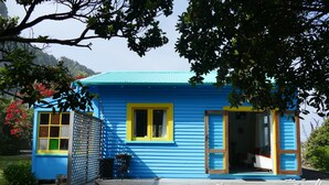 A small, two-story house with a blue exterior and yellow trim. It has a wooden deck and a fenced area with a chair. - Gorgeous Character Bach (Punakaiki)