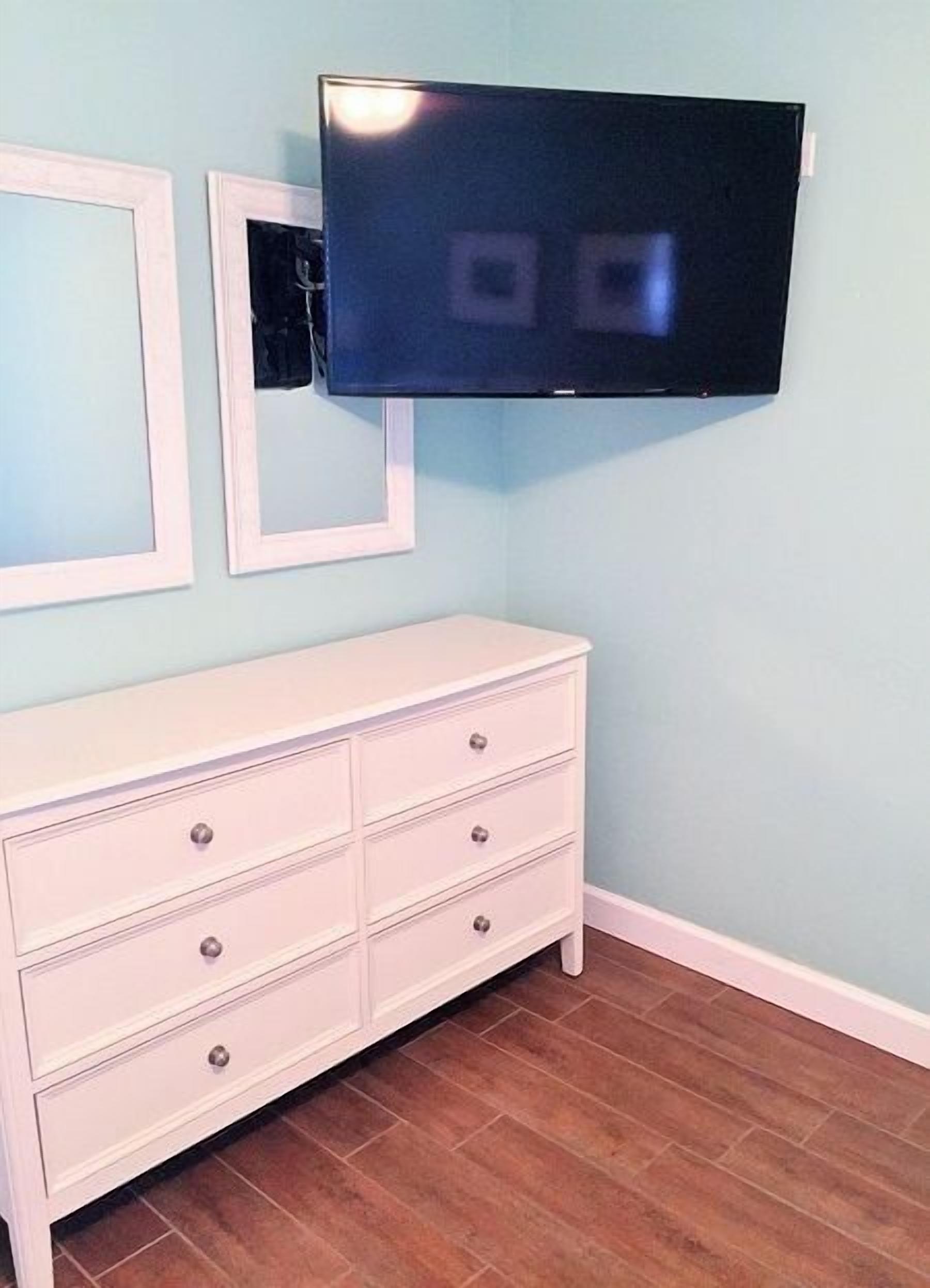 A room with a white dresser, a flat-screen TV mounted on the wall, and a light blue wall.