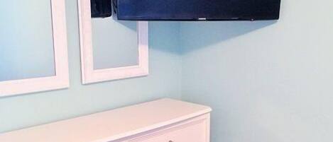 A room with a white dresser, a flat-screen TV mounted on the wall, and a light blue wall.
