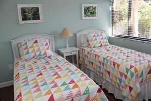 Dos camas individuales con colchas de colores, una mesita de noche con lámpara y una ventana con persianas.
