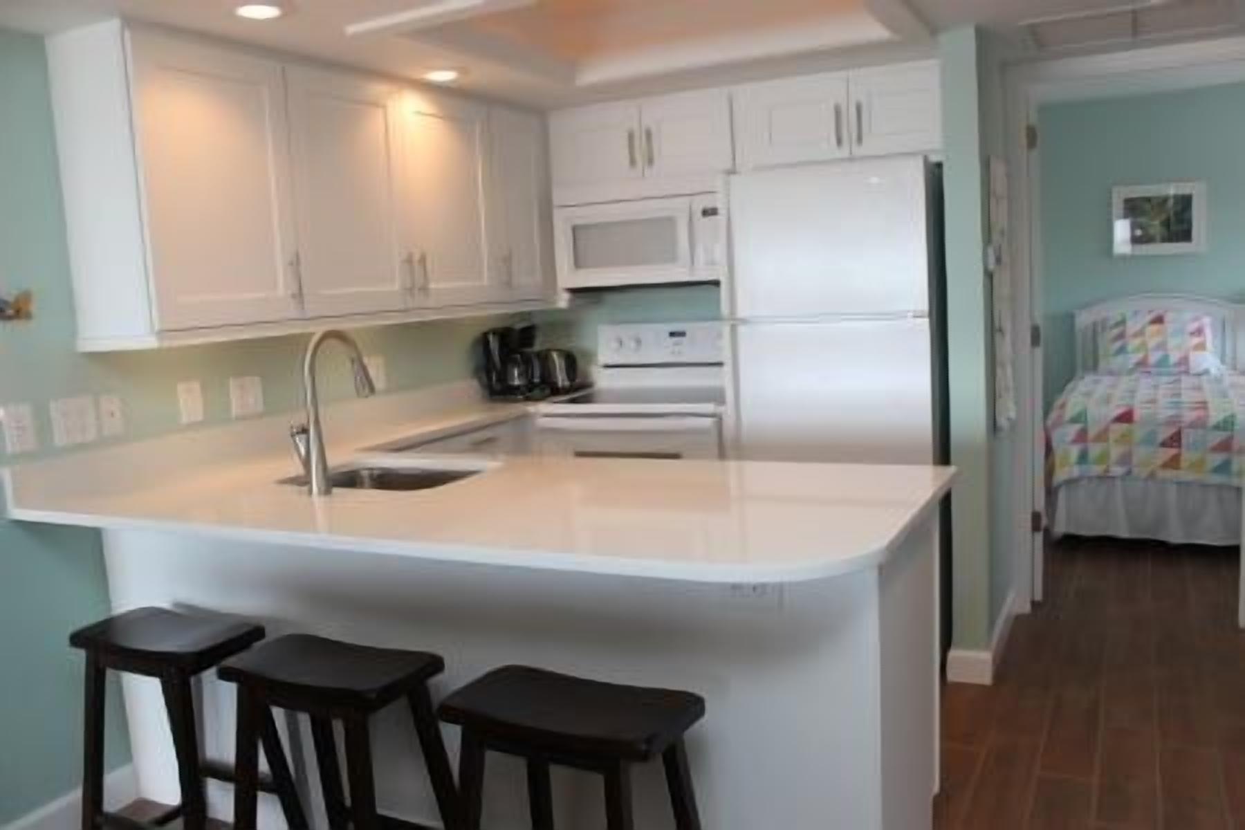 Una cocina con una isla central, armarios blancos, microondas y refrigerador. Hay dos taburetes en la isla y una cama con una colcha colorida al fondo.