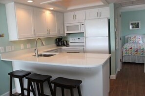 A kitchen with a central island, white cabinets, a microwave, and a refrigerator. There are two stools at the island and a bed with a colorful bedspread in the background. - Firethorn 321- 2 bedroom 2 bath enjoy a fabulous direct beach front view from this 2nd story walk up (Sarasota)
