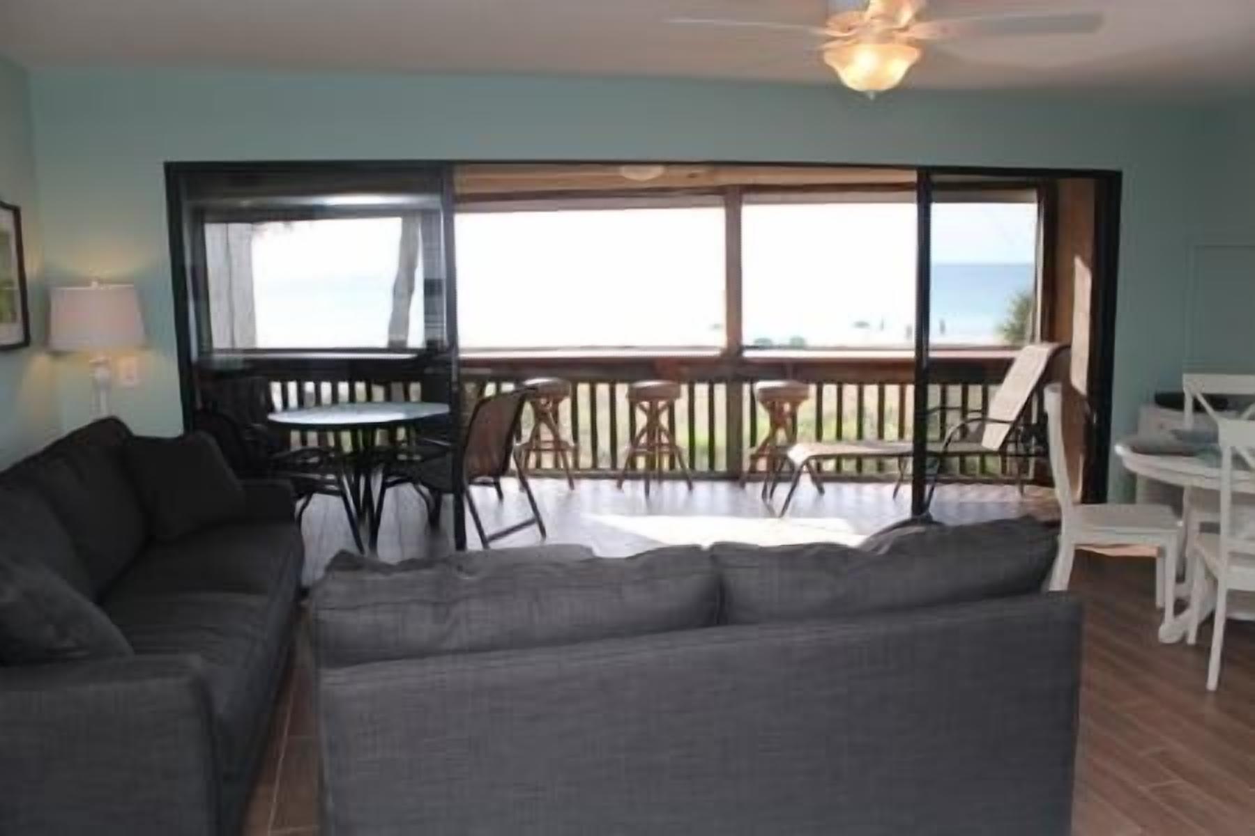 A living room with a sofa, a dining table, and chairs, a balcony with a view of the ocean, and a ceiling fan.