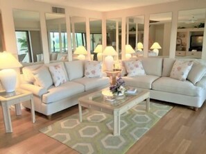 A living room with a sectional sofa, a coffee table, and lamps. There are mirrors on the walls and a rug on the floor.