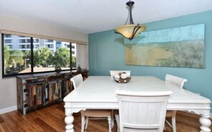 A dining area with a white table and chairs, a decorative cabinet, and a large window with a view of buildings and trees.