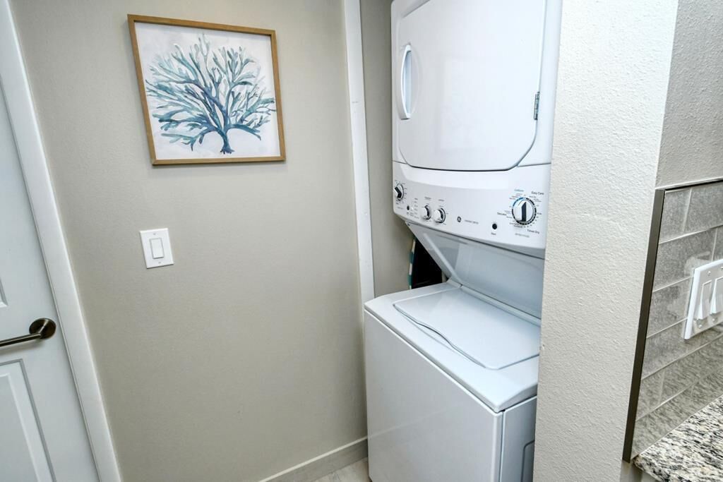 A laundry room with a stacked washer and dryer, a door, and a framed tree artwork on the wall.