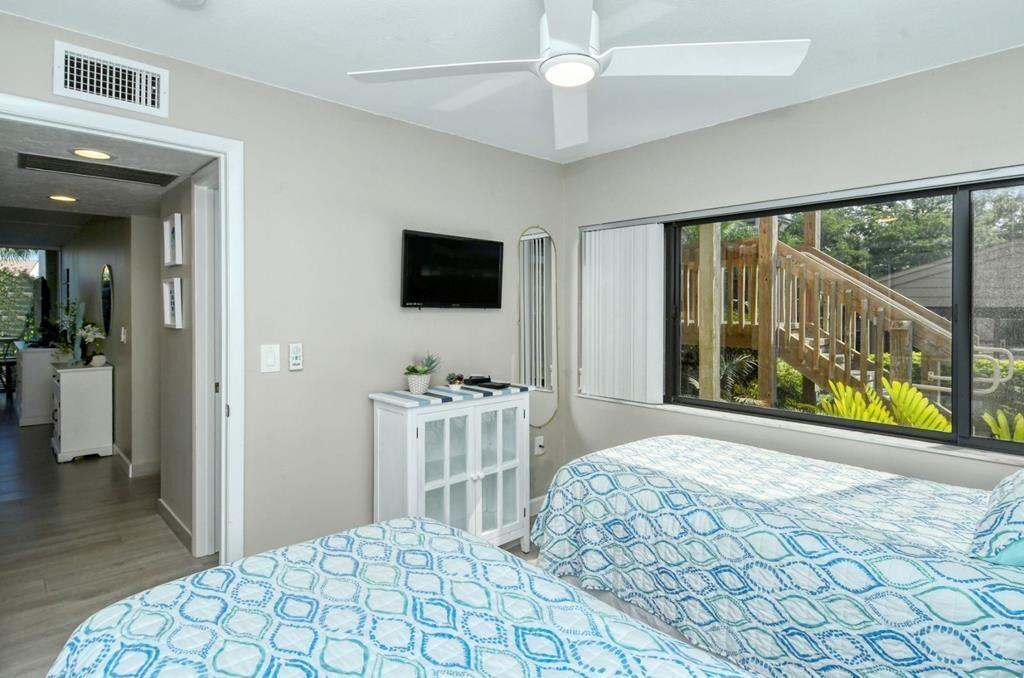 Een slaapkamer met twee bedden, een plafondventilator, een televisie en een raam met uitzicht op een houten terras.