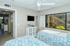 Un dormitorio con dos camas, ventilador de techo, televisor y ventana con vista a una terraza de madera.