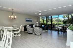 A spacious living room with a dining area, a ceiling fan, and large windows offering a view of the outdoors.