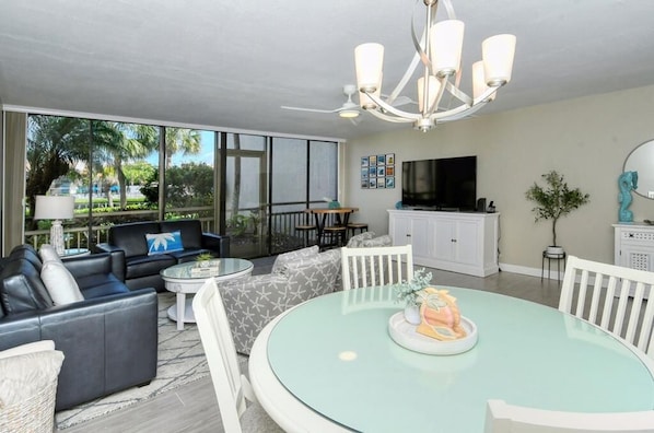 A modern living room with a glass dining table, leather sofas, a flat-screen TV, and a large window offering a view of the outdoors.