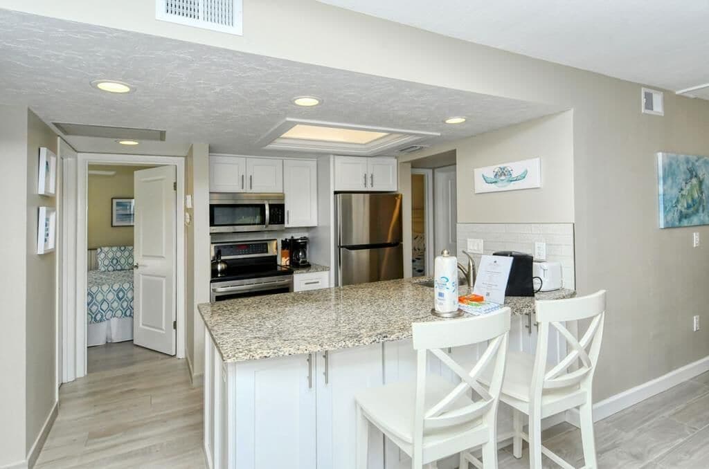 A modern kitchen with white cabinets, granite countertops, stainless steel appliances, and a breakfast bar with white chairs.