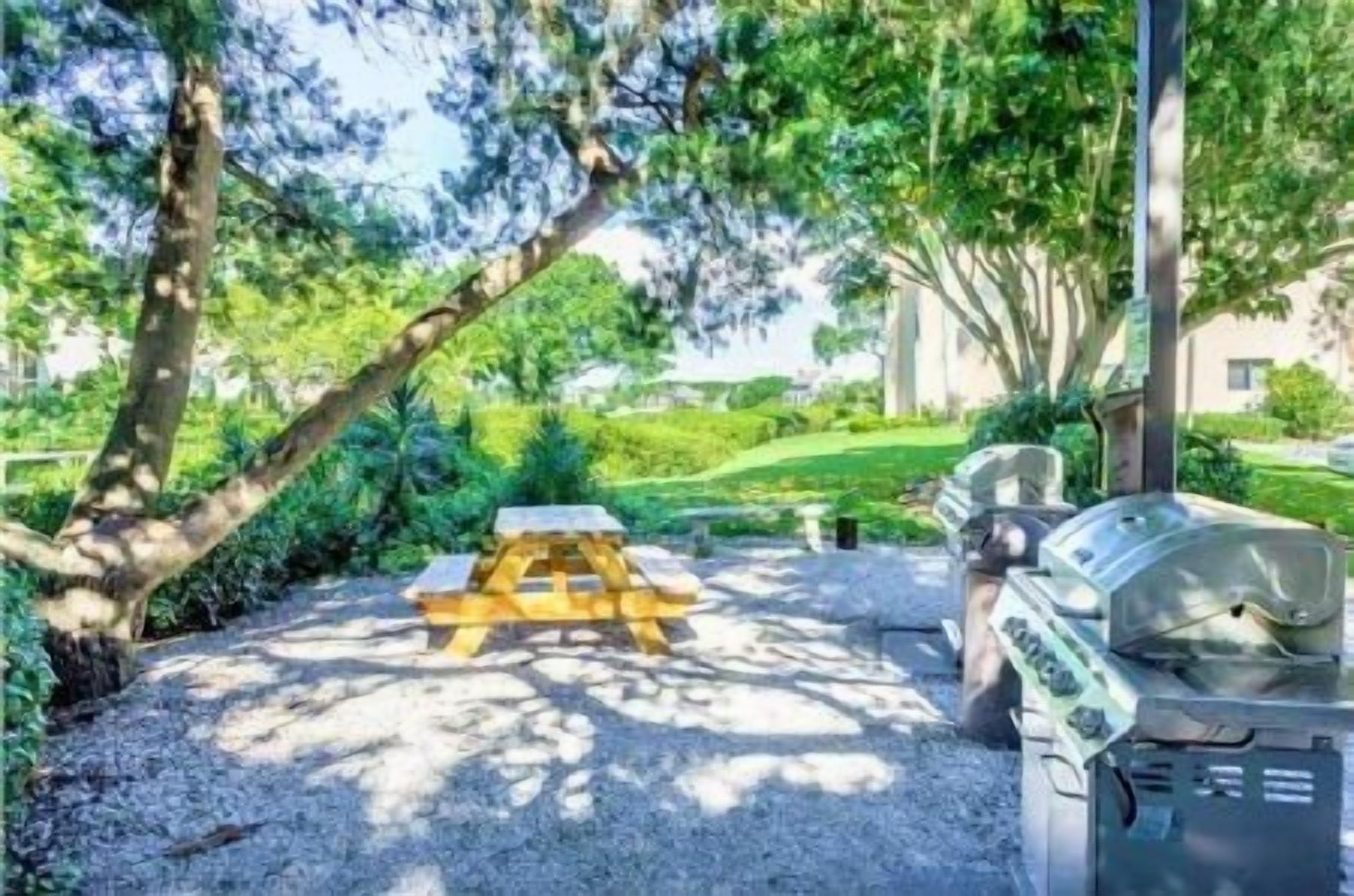 Ein Picknicktisch mit Grill in einer schattigen Außenbereich mit Bäumen und einem Gebäude im Hintergrund.