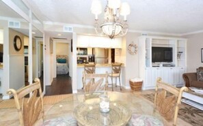 A spacious living room with a round glass table, wicker chairs, a chandelier, and a view into the kitchen area. - Firethorn 613- Ground floor 2 bed / 1.5 bath unit just steps to pool and boat docks (Sarasota)