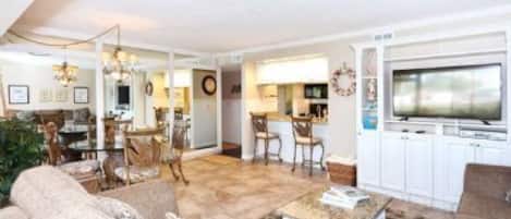 A spacious living room with a large television, a dining area with chairs, and a kitchen area with cabinets.