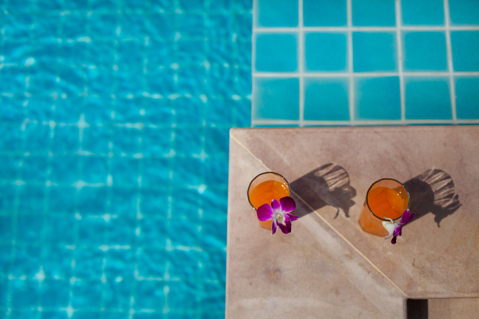 A poolside area with two glasses of orange beverage and purple flowers on a tiled surface.