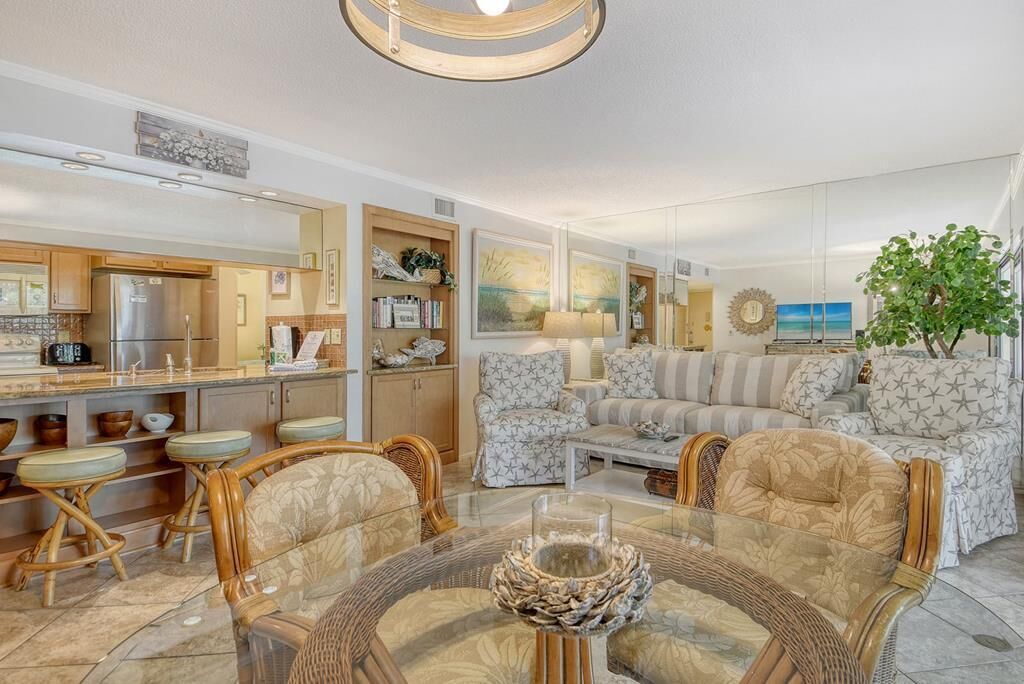A modern living room with a glass coffee table, wicker chairs, a sofa, and a kitchen area with a refrigerator and cabinets.