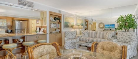 Ein modernes Wohnzimmer mit einem Glastisch, Korbstühlen, einer Couch und einer Küchenzeile mit Kühlschrank und Schränken.