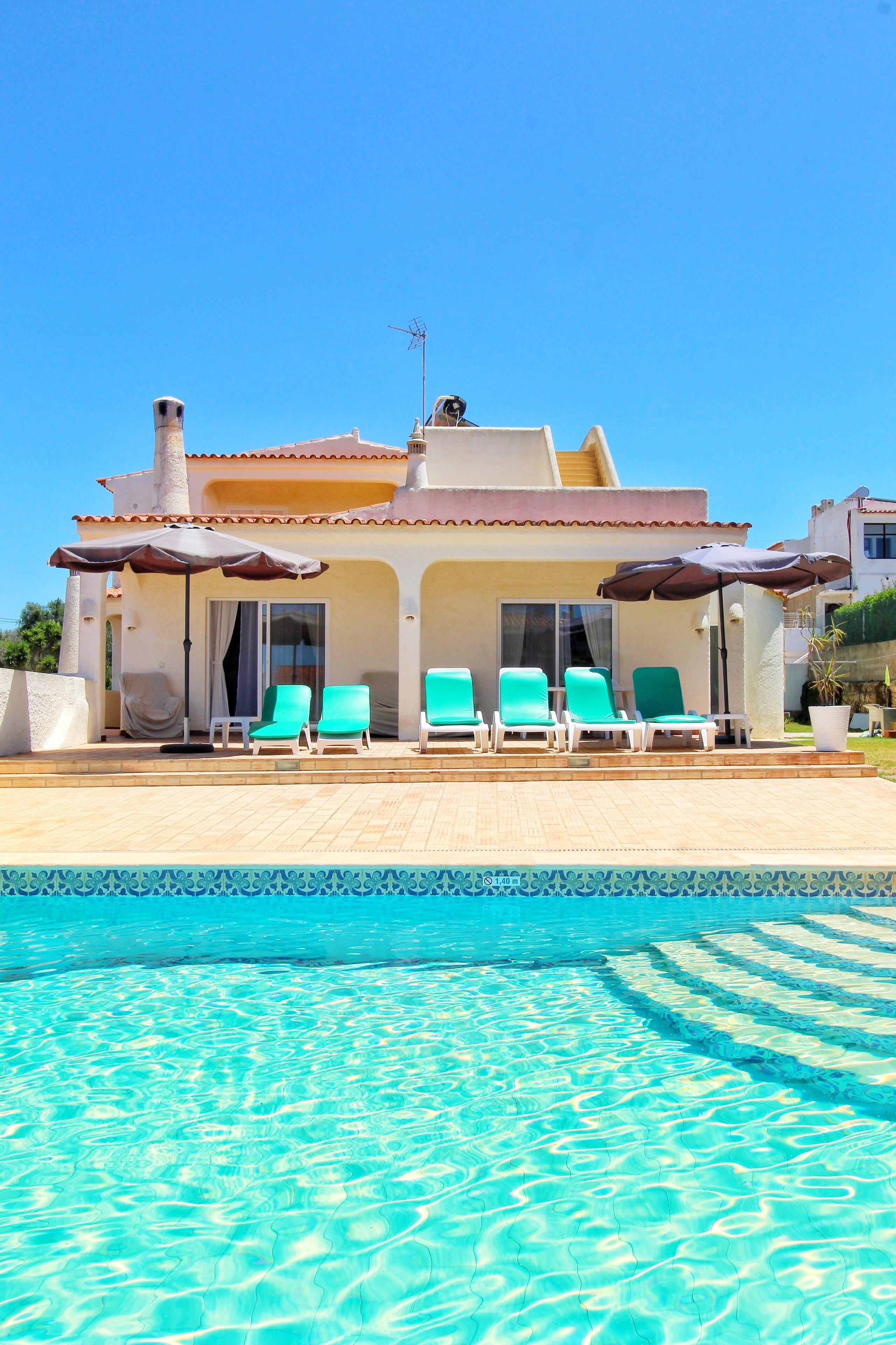 A poolside area with a clear blue pool, a covered seating area with chairs, and a building with a tiled roof.