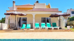 A poolside area with a clear blue pool, a covered seating area with chairs, and a building with a tiled roof. - Portuguese house with a large private pool and very close to the beach. (Carvoeiro)