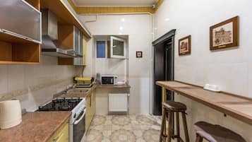 A compact kitchen with a gas stove, microwave, and a small window. There are two stools and a radiator on the floor.