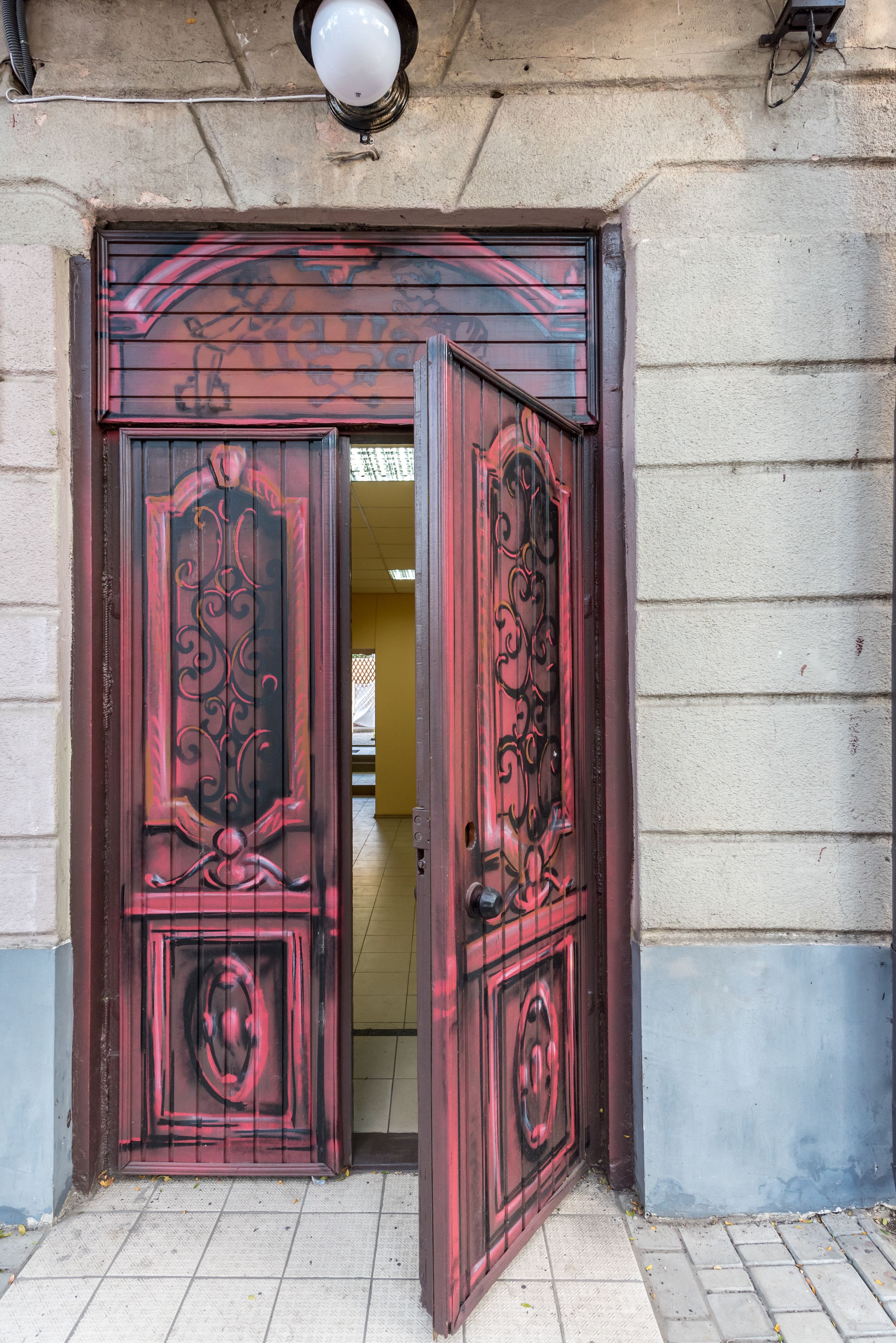 A red door with intricate designs, leading to a building with a tiled floor and a light fixture above.
