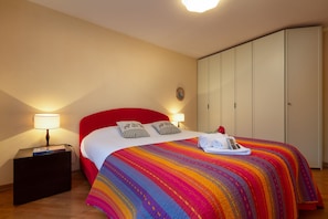 Een net opgemaakt bed met een kleurrijke gestreepte deken, twee bedlampen en een klein nachtkastje met boeken.