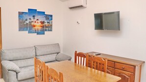 Ett vardagsrum med en grå soffa, ett matbord i trä och stolar. På väggarna hänger tre tavlor som föreställer tropiska motiv. En platt-TV är monterad på väggen ovanför bordet.