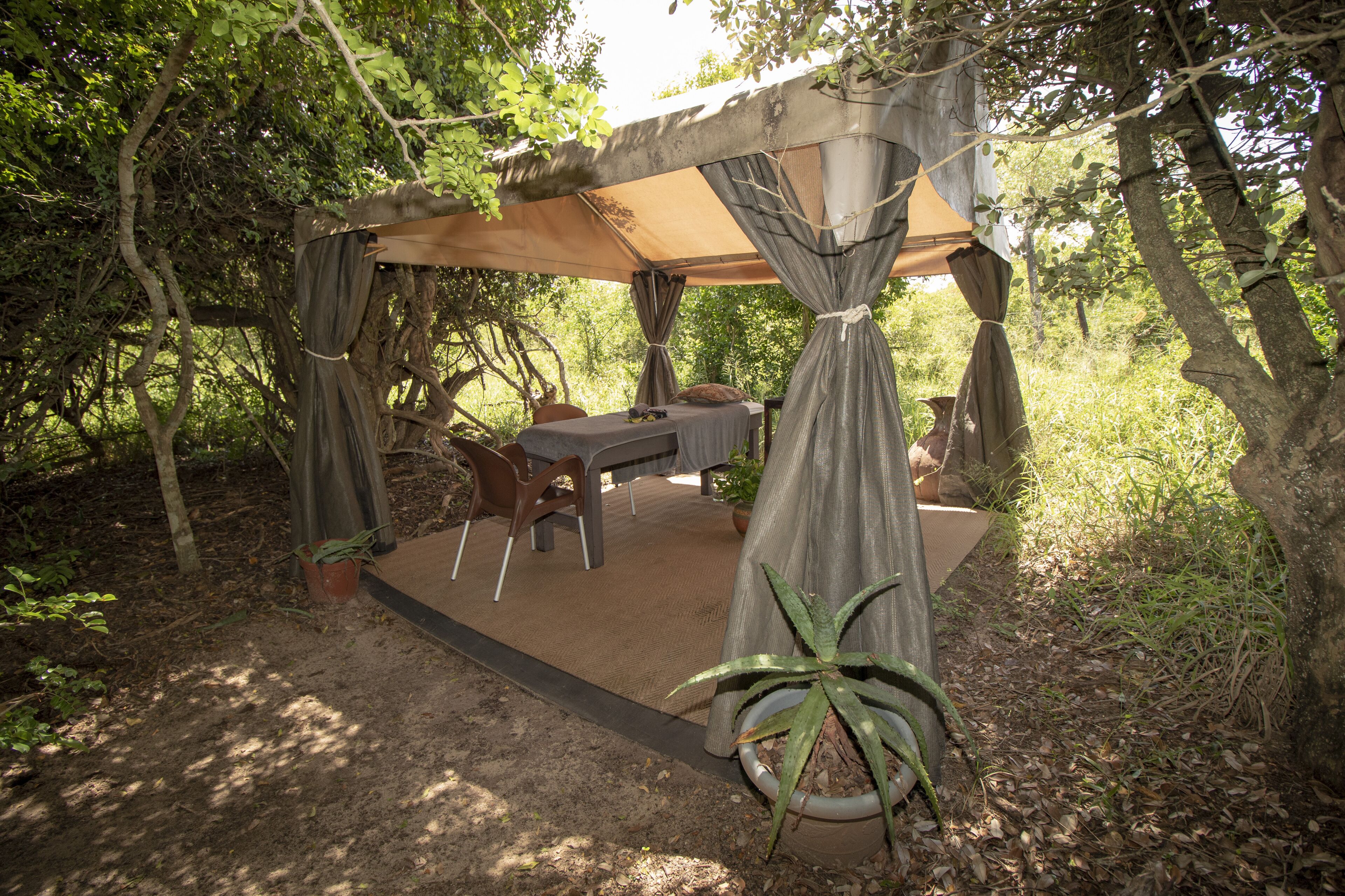 A covered outdoor dining area with a table and chairs, surrounded by trees and potted plants.