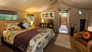 A tented room with a bed, a small table with a lamp, and a wicker chair.