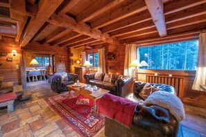 A log cabin interior with a dining area, two sofas, a fireplace, and a large window with a view of the forest.
