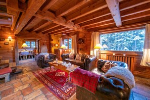 L’intérieur d’une cabane en rondins, avec une cheminée, des canapés en cuir, un coin repas et une vue sur une forêt enneigée.