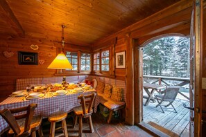 L’intérieur chaleureux d’un chalet avec une table à manger en bois dressée pour un repas, un coin salon avec des bancs et une vue sur une forêt enneigée par une grande fenêtre.
