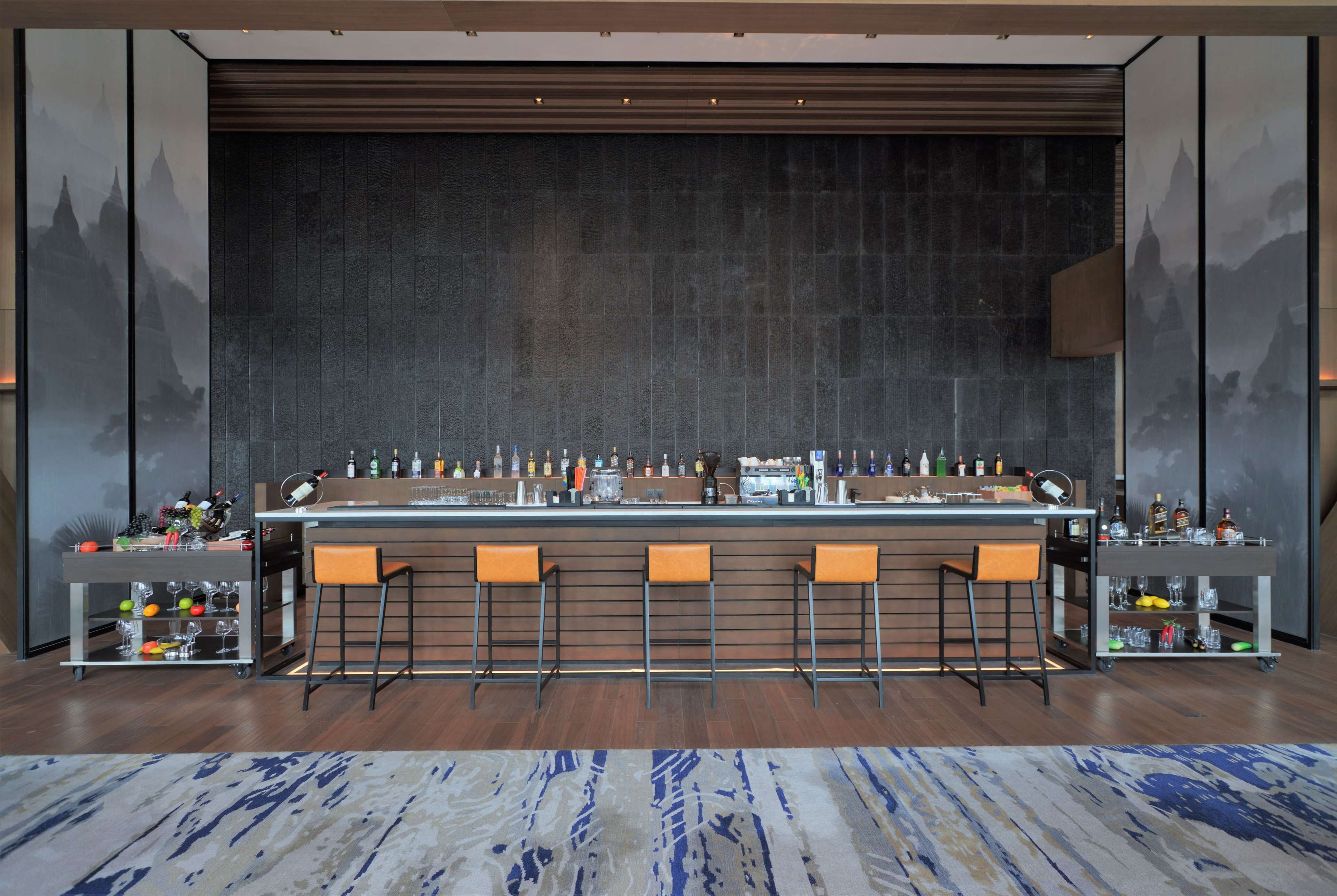 A bar area with a long counter, multiple stools, and shelves with bottles and glasses.