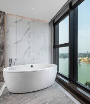 Um banheiro moderno com uma banheira independente, azulejos de mármore e uma janela grande que oferece vista para a paisagem urbana e um corpo d'água.
