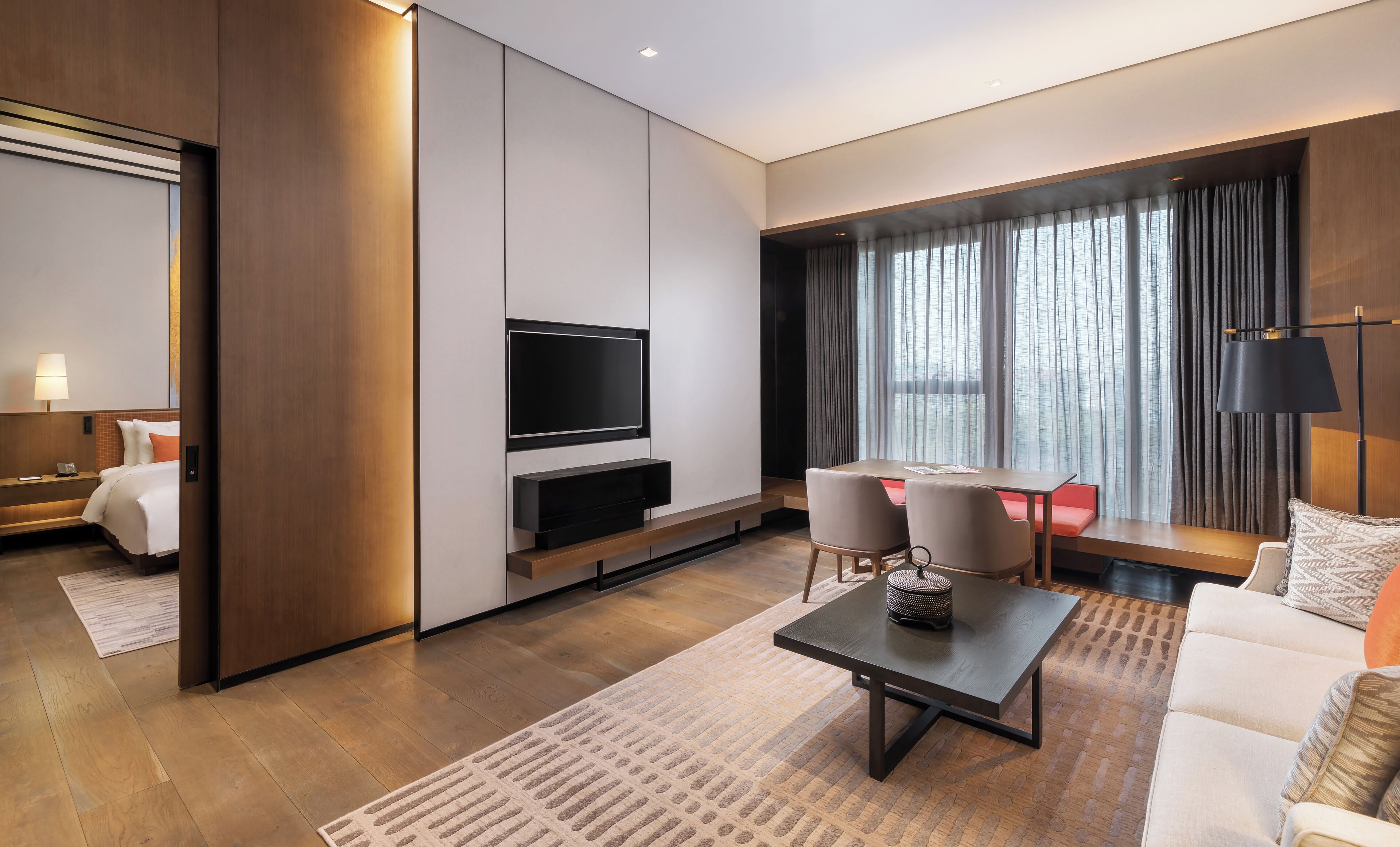 Ein modernes Hotelzimmer mit einem Flachbildfernseher, einer Essecke mit Tisch und Stühlen, einem Sofa und einem großen Fenster mit Vorhängen.