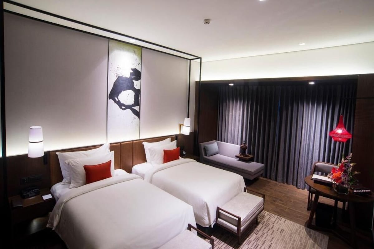 A hotel room with two beds, a sofa, a desk, and a large window with curtains.