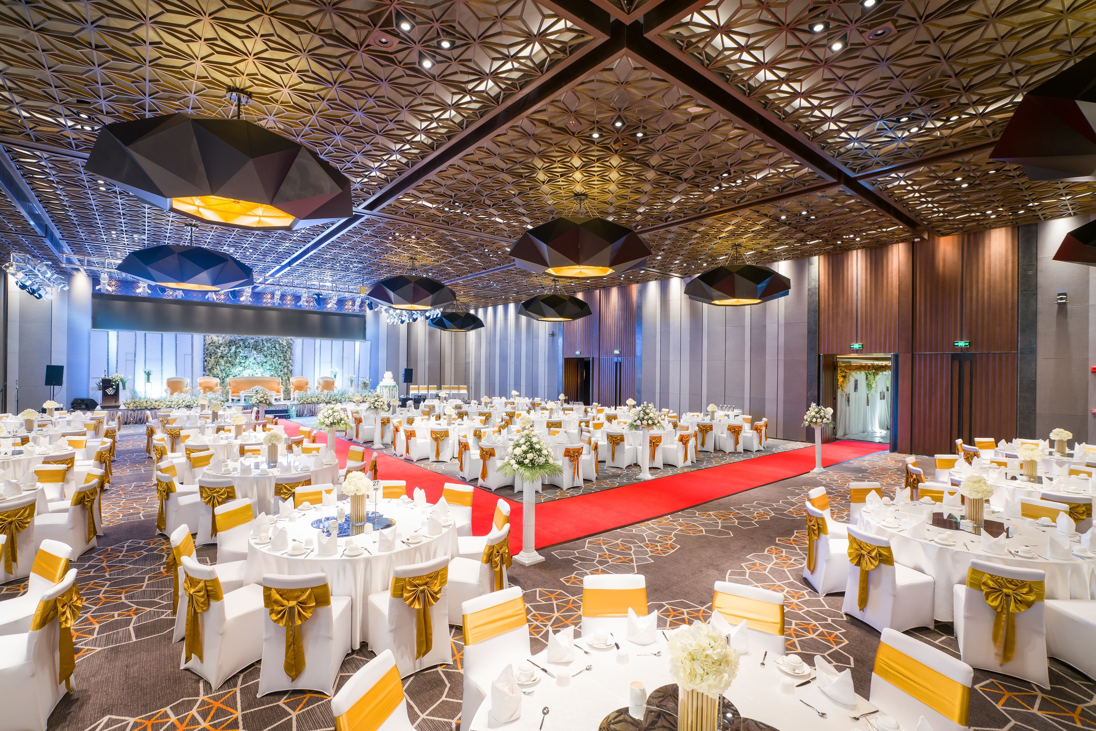 A banquet hall with round tables, white chairs, and gold sashes, featuring a geometric ceiling design and a red carpet.